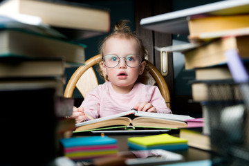 A little girl in mother s glasses sits behind books and notebooks