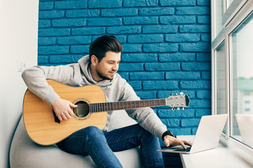 handsome man with guitar using his laptop at home with the wall of blue bricks
