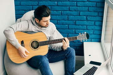 Obraz premium cheerful guitarist playing on instrument at home with laptop and phone on the side