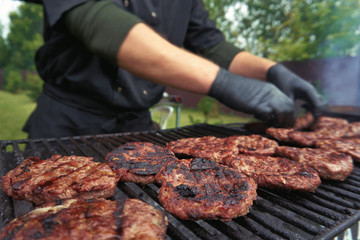 Close up of grilled beef burgers. Lot of burgers on grill. Hands of cook in black protective gloves touching beef patty. Cook working outdoor. Open air grill station. Fast food festival. Summer party