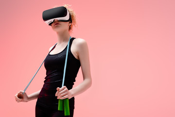 young woman in in sportswear and virtual reality headset holding jumping rope on pink