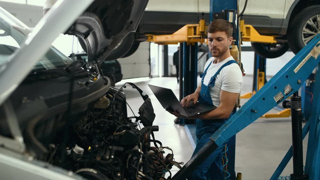 Handsome mechanic in uniform is using a laptop while repairing car in auto service.