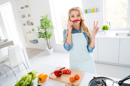 Photo of cheerful housewife blond lady cut salad cooking tasty dinner breakfast hold red hot chilli pepper like mustache showing v-sign symbol playful good mood stand modern kitchen indoors