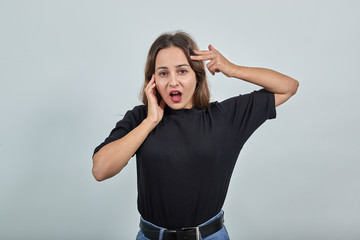 Cute young woman brunette in black t-shirt, blue jeans with belt on gray background surprised, she opened her mouth, held cheek, and put hand to head