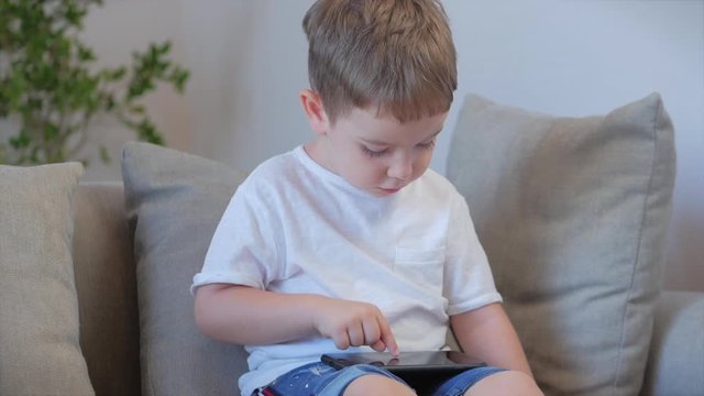 Curious cute boy preschool child using a digital tablet technology device looks telephone.Plays in the phone sitting on the sofa alone.Children tech addiction concept play game at home.