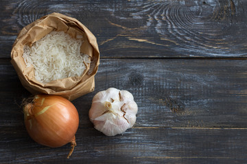 Raw white long-grain basmati rice in a craft bag with onions and garlic on a wooden table, ingredients for a delicious vegan dish, top view, space