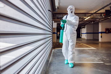 Full length of worker in sterile protective suit and mask sterilizing door of a garage from corona...