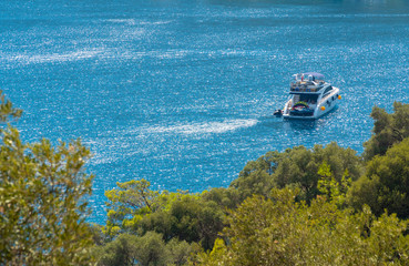 A white luxury yacht with turkish flag and orange boat fender anchored in shimmering blue Aegean waters. View on a sunny calm summer day from land behind green trees