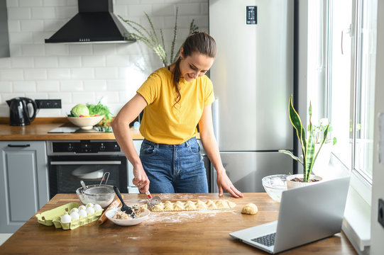 A young woman cooks at home in the kitchen, she watches a video recipe on a laptop and works with the dough on the table, she cuts ravioli. Italian cuisine at home - Powered by Adobe