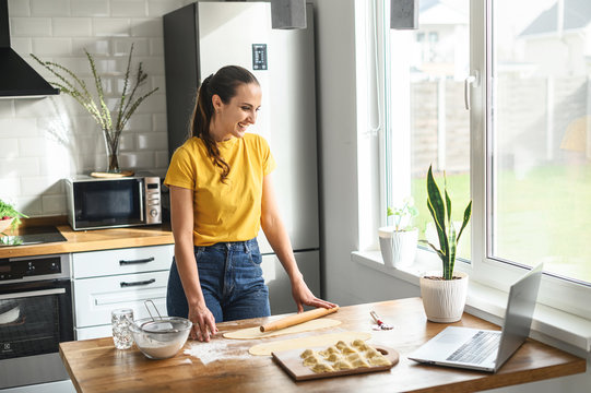 Cooking at home. Positive girl watching online video recipes on a laptop and cooking in the kitchen at home. She rolls dough with a rolling pin