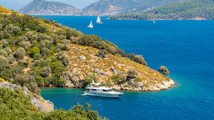 Fototapeta na wymiar Summer concept: Luxury white yacht boat anchored stern in a bay with blue turquoise waters. View from land with sky, sea and sailing boats in background with copy space