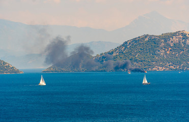 Dark smoke covering the air of a boat on fire at sea. Blue waters with sailing boats, islands, sky...