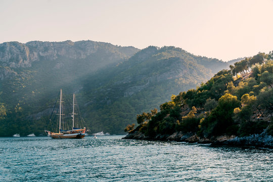 Summer Concept: A Turkish Gulet And Behind Some Luxury White Yachts Anchored At The Aegean Sea With Sun Beam In Background. Natural Photo With Copy Space. Green And Blue Contrast. No People