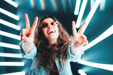 Cheerful cute smiling girl shows peace sign in round glasses at neon background.