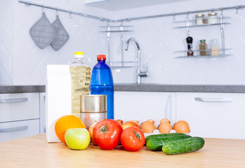 Set of fresh raw vegetables tomato, cucumber, egg, orange, apple, bottle of water and oil, milk on a wood table in a modern kitchen room.