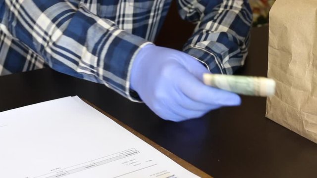 A man in rubber gloves recounts dollars and hands them to the courier per frame. Nearby are paper bags with food and a delivery form.
