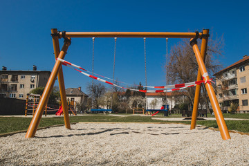 Closed children playground or swing on a chain due to corona virus restrictions taking in effect. Red and white tape over children swing.
