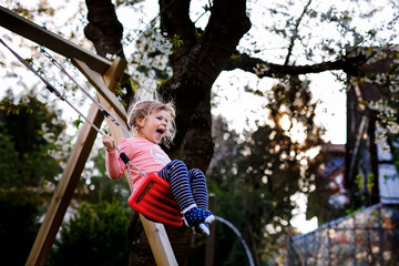 Happy beautiful little toddler girl having fun on swing in domestic garden. Cute healthy child swinging under blooming trees on sunny spring day. Baby laughing and crying