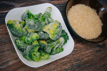 Frozen green cabbage, semi-finished product in a white cup. On a dark wood table. Nearby is a dark cup with white rice. Recipe