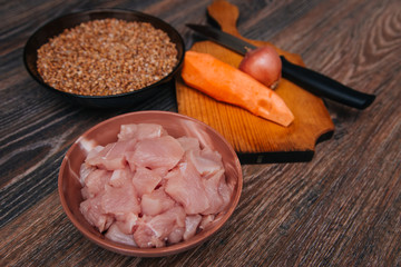 Ingredient for cooking carrots and onions on a blackboard. Uncooked diced turkey in a black cup on a dark kitchen table. Cut the fillet into pieces. Chicken meat. A cup of buckwheat.