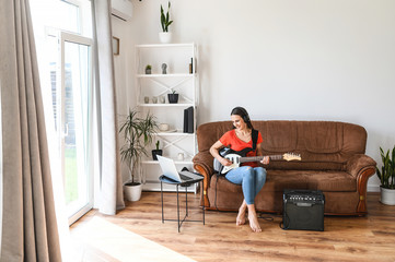 Fototapeta na wymiar Online learning concept. Young woman watching video tutorials and learning to play the electric guitar, uses a combo amplifier