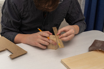 a teenager in a work coat sits at a desk draws a drawing on a box for cutting wood. activities at home during the period of quarantine and self-isolation