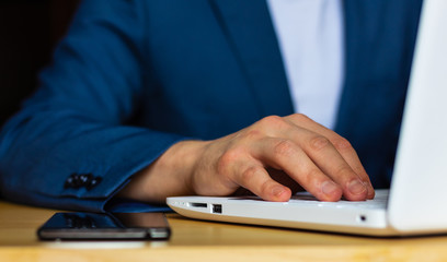 Close-up of the hands of a well-dressed man in a blue suit and a white shirt using a laptop at a desk. With a smartphone.