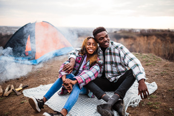 African American man and woman sitting on rug near campfire and tent, smiling, hugging each other....