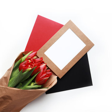 Red tulips bouquet in craft paper with craft photo frame with blank white space and two envelopes for greeting cards. White background, flat lay.