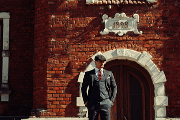 Portrait of retro 1920s english arabian business man wearing dark suit, tie and flat cap standing against old brick house 1898 year.