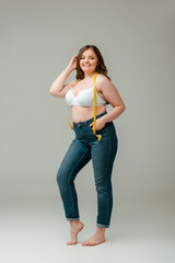 cheerful plus size woman in jeans and bra standing with measuring tape on grey