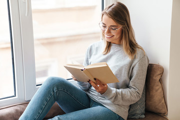 Photo of smiling woman reading book while sitting near window