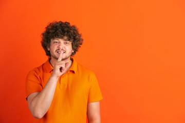 Fototapeta na wymiar Whispering a secret. Caucasian young man's monochrome portrait on orange studio background. Beautiful male curly model in casual style. Concept of human emotions, facial expression, sales, ad.