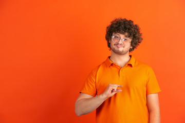 Showing something little. Caucasian young man's monochrome portrait on orange studio background. Beautiful male curly model in casual style. Concept of human emotions, facial expression, sales, ad.