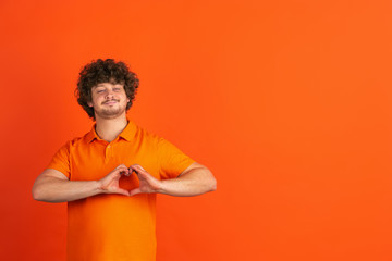 Heart gesture. Caucasian young man's monochrome portrait on orange studio background. Beautiful male curly model in casual style. Concept of human emotions, facial expression, sales, ad.