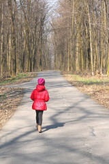 Little girl in red jacket and red hat walks in spring forest among trees on asphalt road