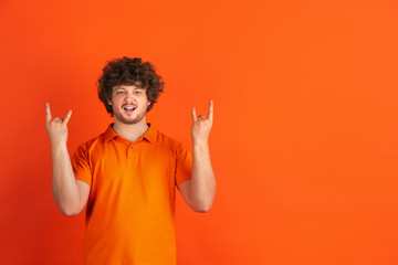 Rock horn gesture. Caucasian young man's monochrome portrait on orange studio background. Beautiful male curly model in casual style. Concept of human emotions, facial expression, sales, ad.