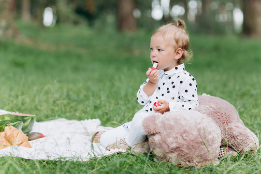 Pretty little girl is playing with teddy bear toy outdoors and doing make up with lipstick, cute child having fun in the park on picnic in summer time, happy childhood concept