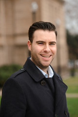 Hispanic businessman standing against a wall in a black coat. The man smiles openly.