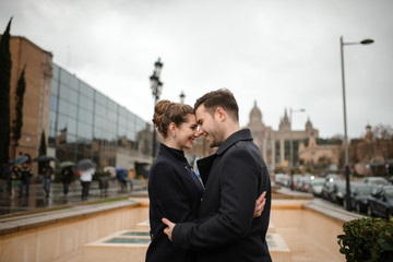 Young beautiful loving Hispanic couple walks under an umbrella during the rain in Plaza Spain. Couple posing against the backdrop of the National Museum of Art of Catalonia.