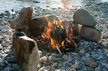 Near the river campfire ablaze with stone circle. Camping season, summer, joy.