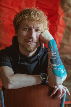 portrait of redhead stylish man with tattoo on his hand sitting on the chair on red background. Calm, fashion, tattoo, style concept. Film grain