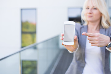Mockup blonde woman holding her smartphone with empty screen in a modern building. Real Estate Agent with mockup smartphone. Young woman with phone in modern background