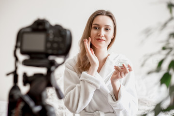 A yound woman blogger sits in her room in a white bathrobe holds a jar bottle of a cream and puts...