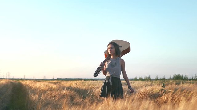 Young hipster girl holding guitar in hand and walking in the wheat field. Woman with acoustic instrument enjoying nature and sunlight. People, lifestyle, student, expression and style concept.