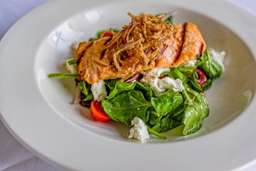 Stacked grilled salmon with onion rings salad, vegetable, and sauce on white plate