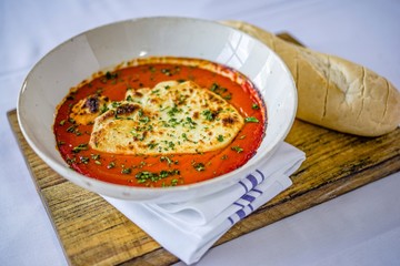 Grilled mozzarella cheese with roasted red pepper and tomato sauce, sprinkled parsley, and french bread baguette in a white bowl on a wooden cutting board.