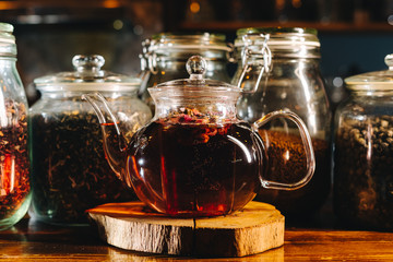 A teapot of black tea standing in front of jars filled with tea leaves, warm light, close up