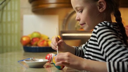Beautiful blond child girl in striped dress hand painting easter egg with paint at kitchen table with fruit plate at background. Cosy home wooden kitchen