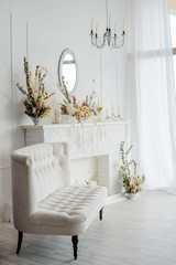 Elegant home interior in white tones with vintage couch, crystal chandelier, mirror under fireplace decorated dried flower comosition.